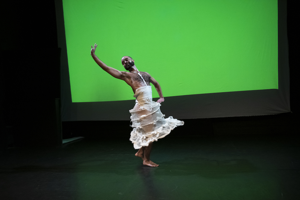 on a black stage with a lime green background, a bearded black male is caught in an exuberant waving pose..his hairy chest is exposed and he wears a modern modified version of a netted, hoop skirt